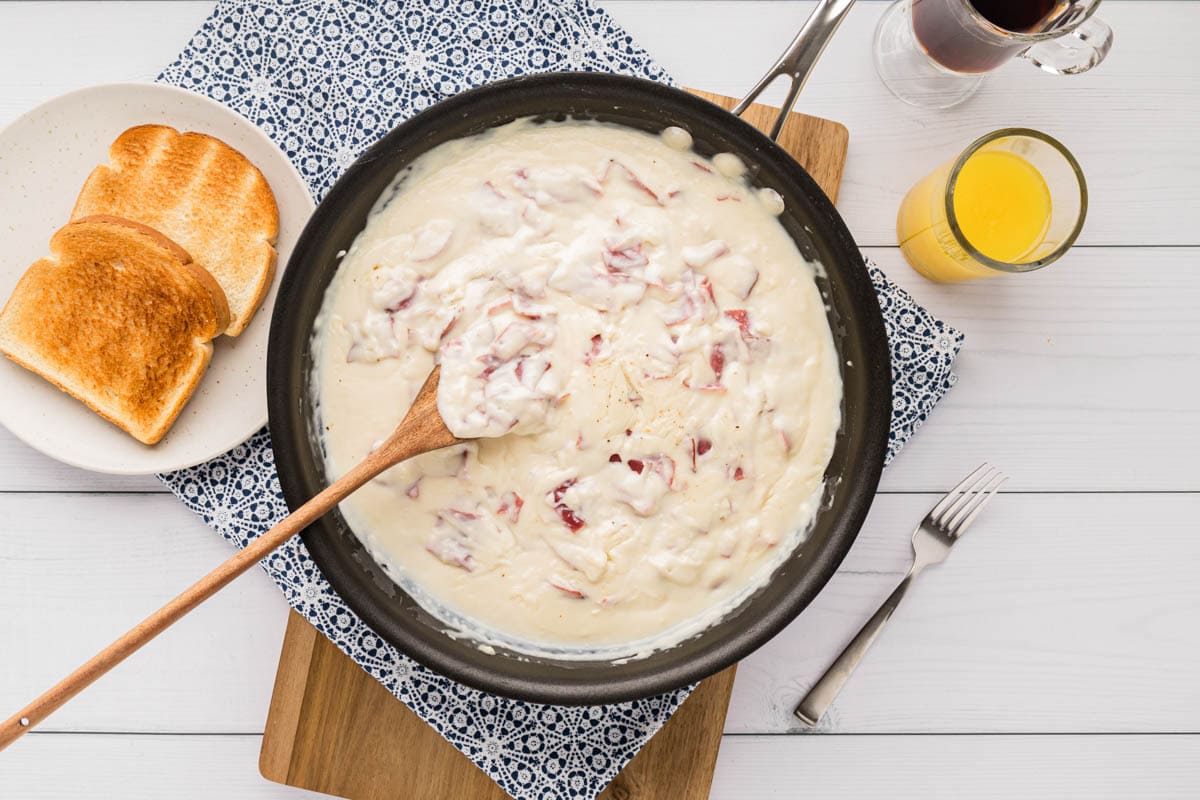 A skilled filled with creamed chipped beef gravy set on a white wood table with slices of toast on a serving plate and coffee and juice.