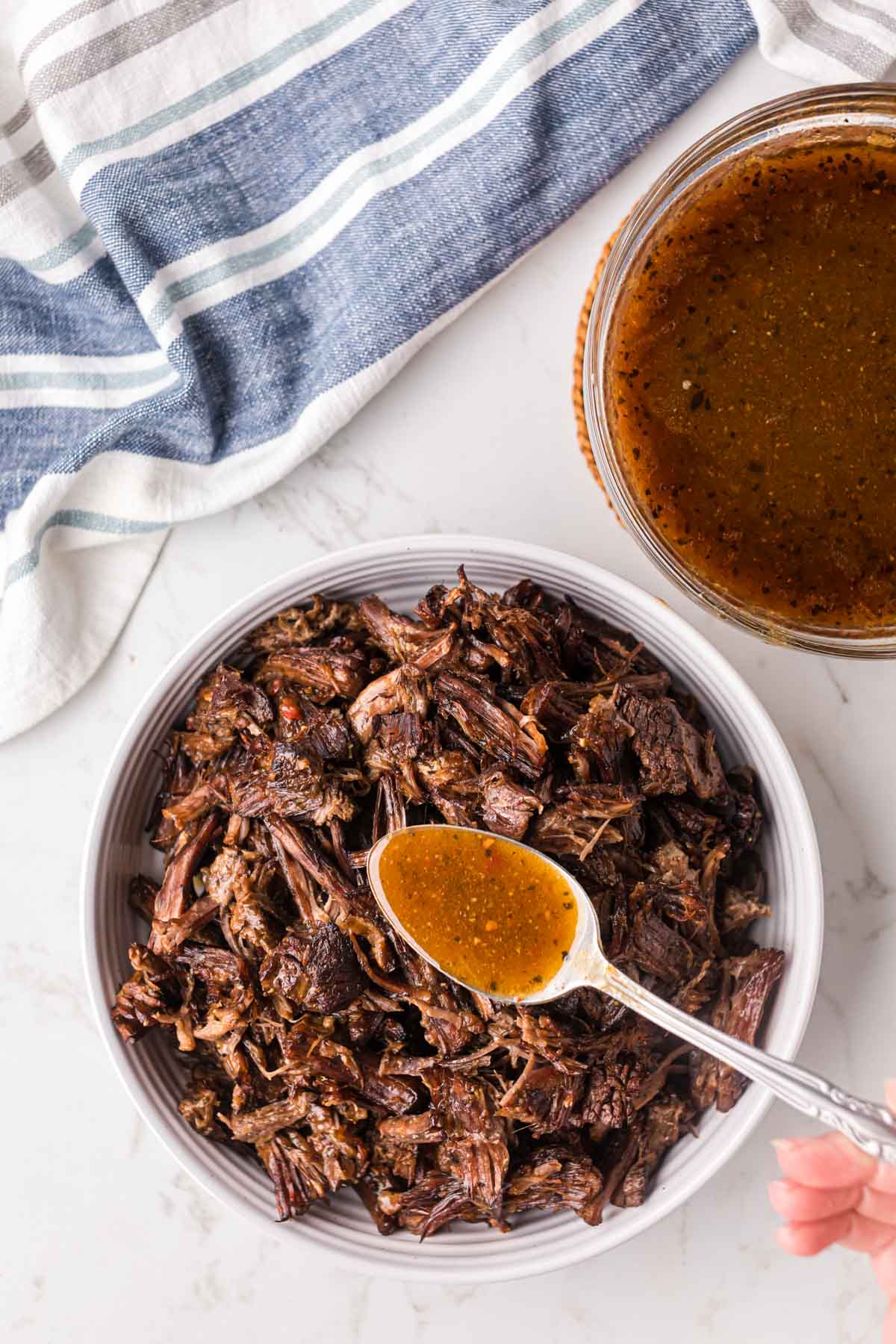 A serving bowl of shredded beef Barbacoa with a spoonful of cooking liquid being poured over the top.