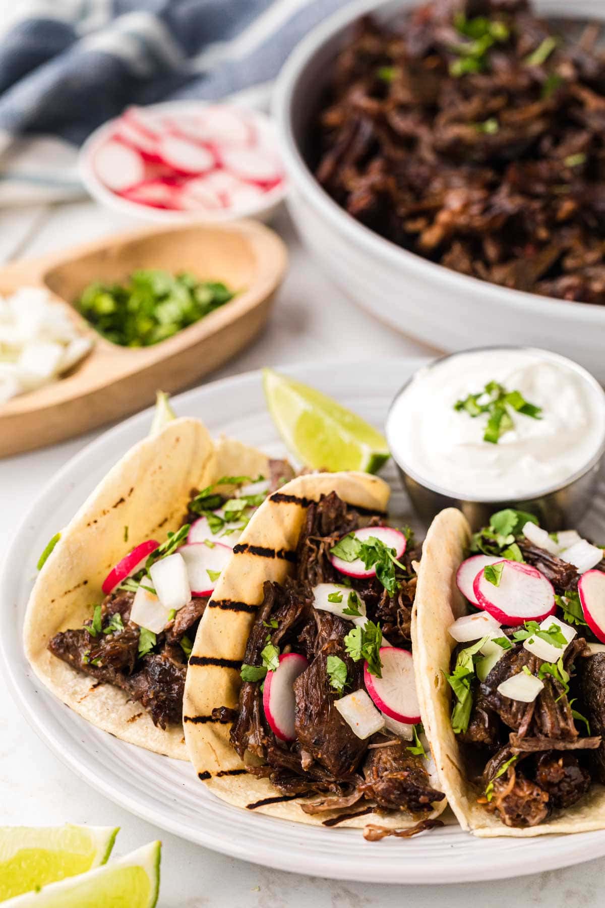 Three beef barbacoa tacos on a white serving plate with lime crema and a slice of lime on the side.