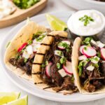 Three beef barbacoa tacos on a white serving plate with lime crema and a slice of lime on the side.