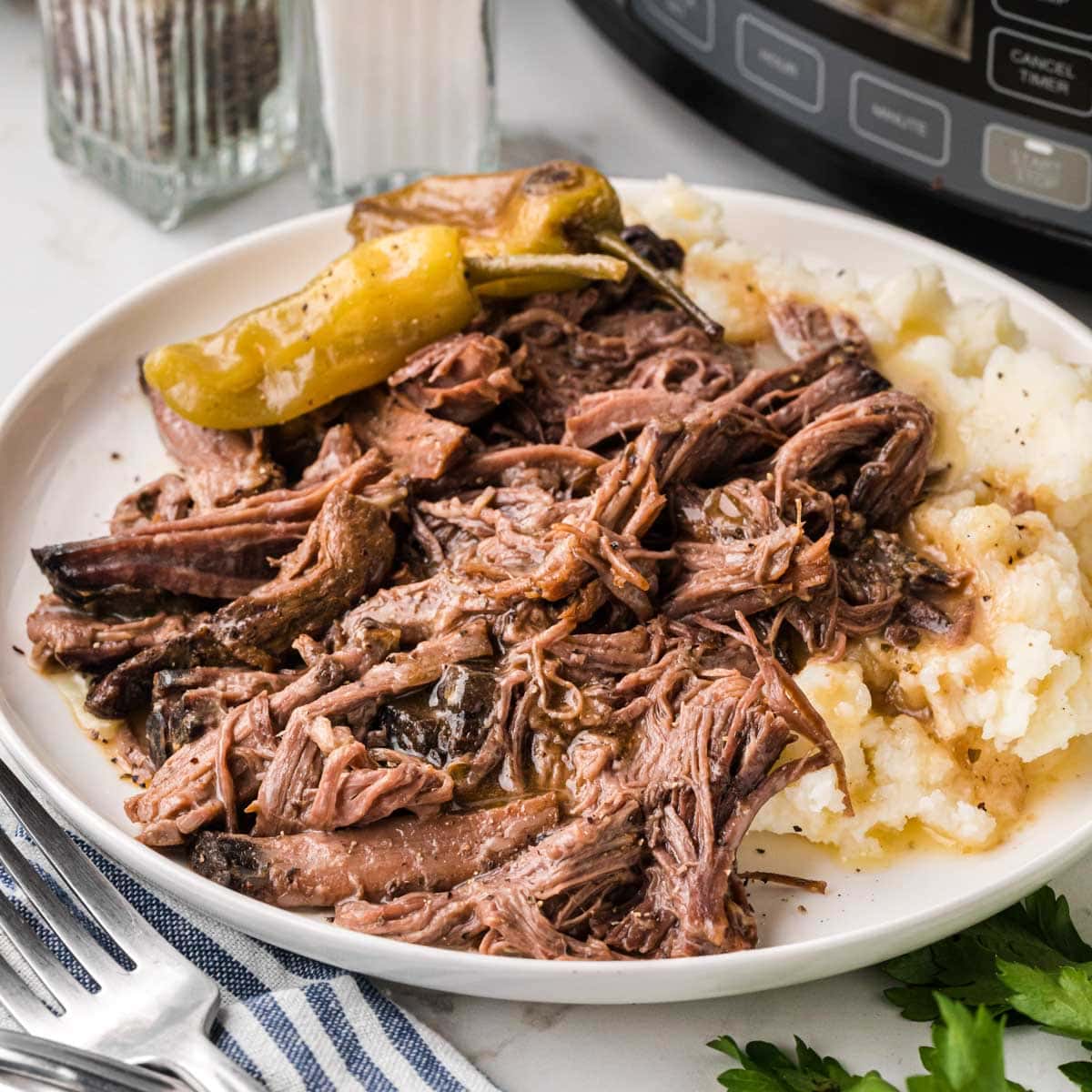 A white plate with a bed of creamy mashed potatoes topped with shredded Mississippi pot roast.