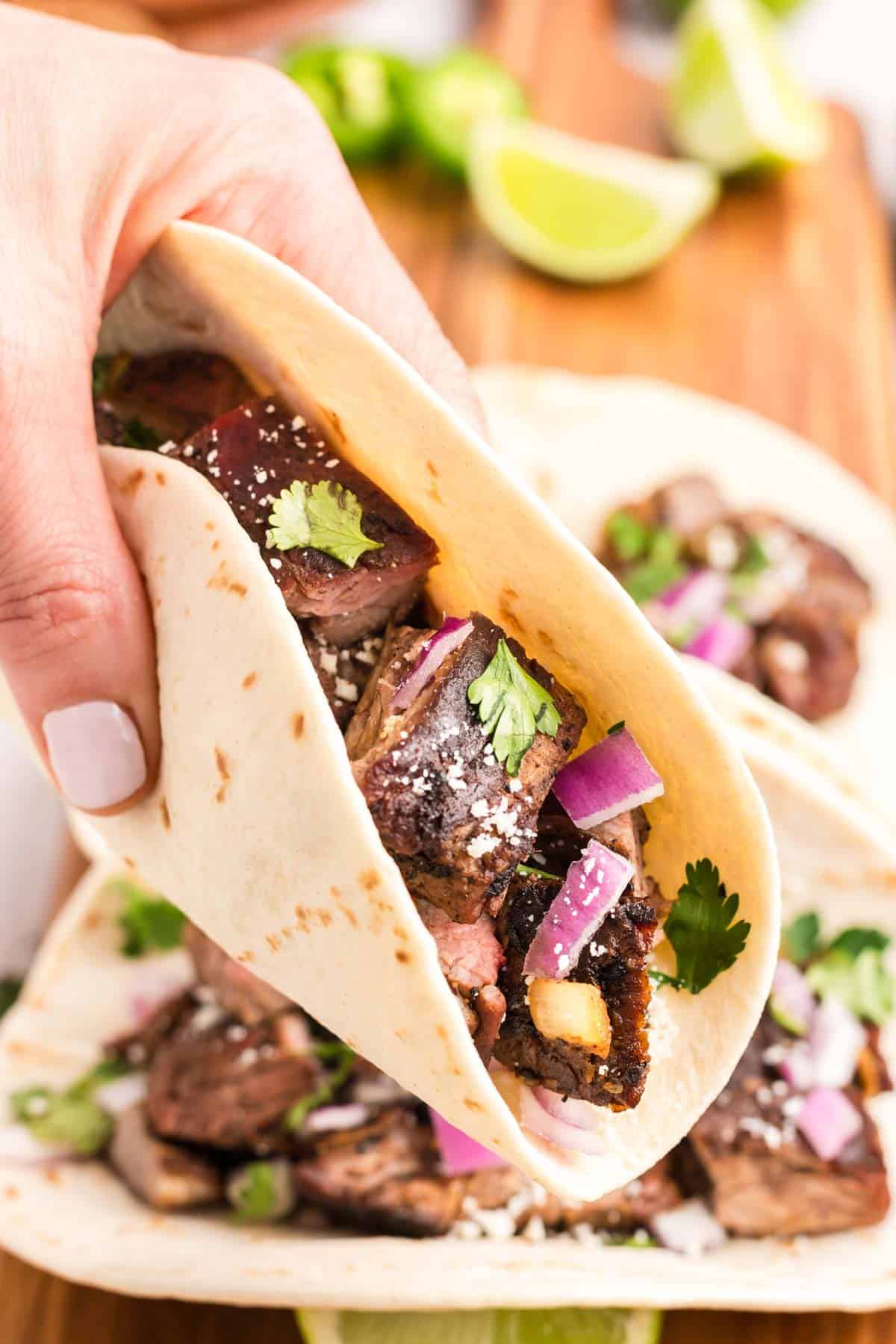A hand picking up a carne asada street taco garnished with onion, cilantro and cotija cheese.