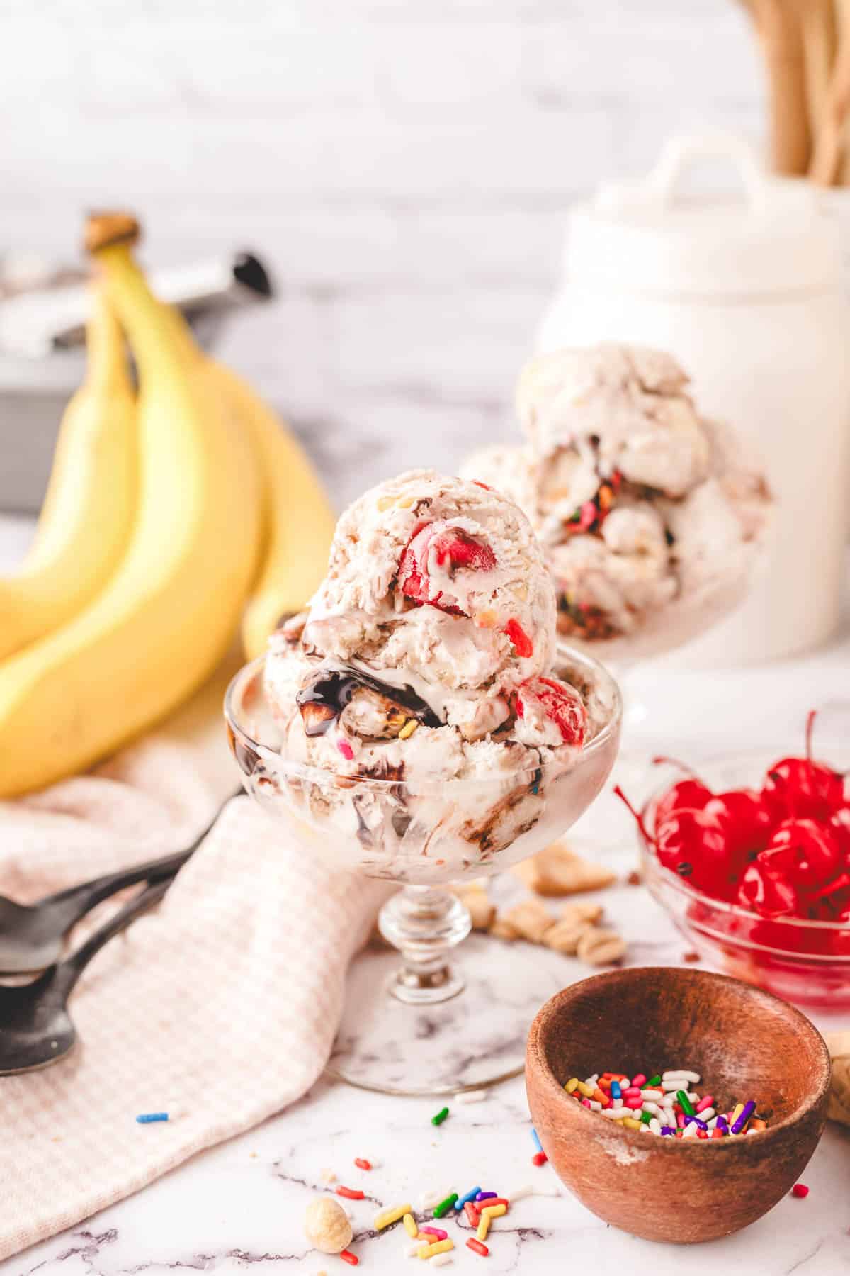 A glass dessert bowl with scoops of banana slip ice cream with sprinkles, cherries, and banana condiments.