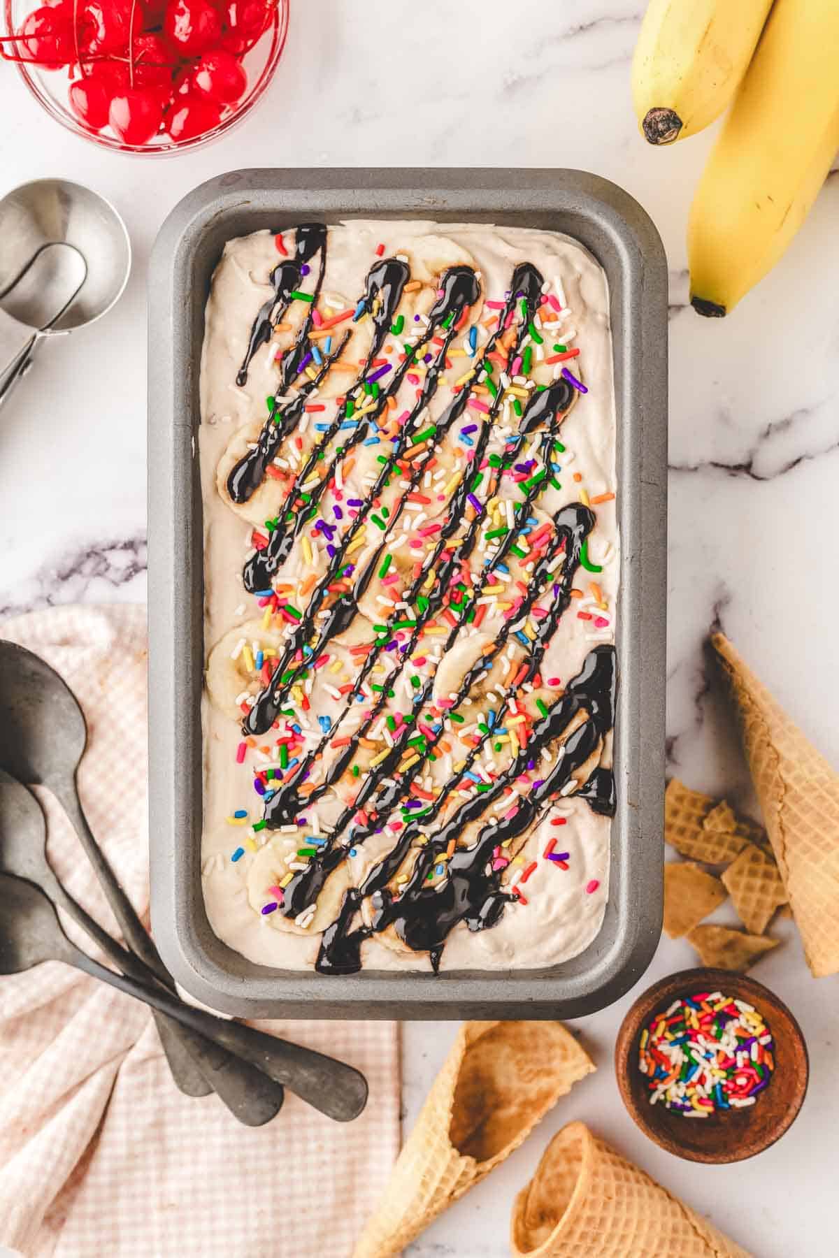 A loaf pan filled with no-churn banana split ice cream topped with sprinkles and drizzled with chocolate syruo