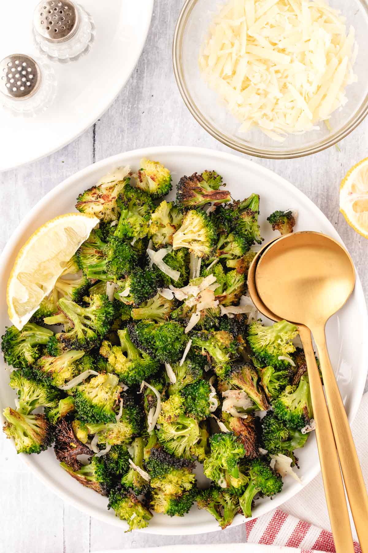 A serving bowl of broccoli that has been roasted until golden brown with parmesan cheese and served with a wedge of lemon.