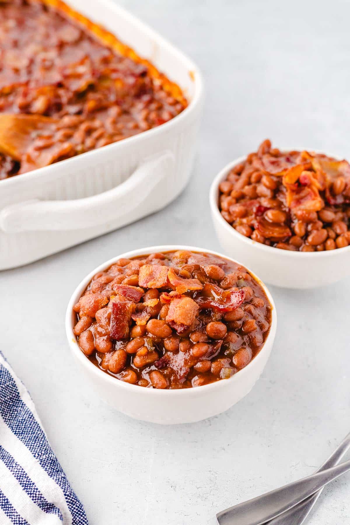 Two bowls of baked beans with casserole serving dish in background.