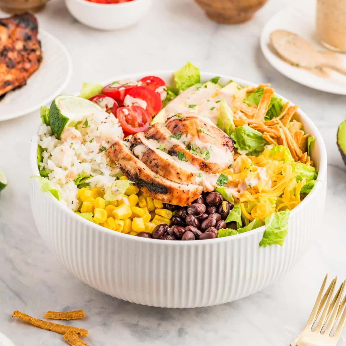 A white serving bowl with a bed of lettuce greens topped with bbq chicken, rice, cheese, corn, black beans, avocado, tomatoes and topped with a bbq lime ranch dressing.