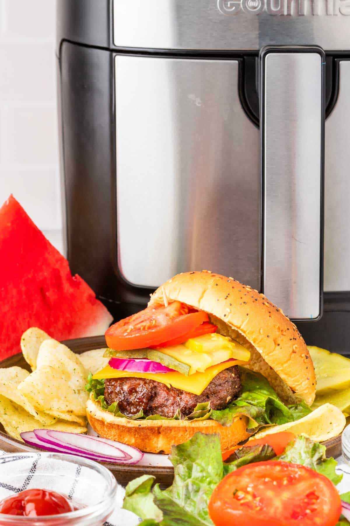 A cheeseburger on a bun with condiments on a plate along with chip and an air fryer in the background.