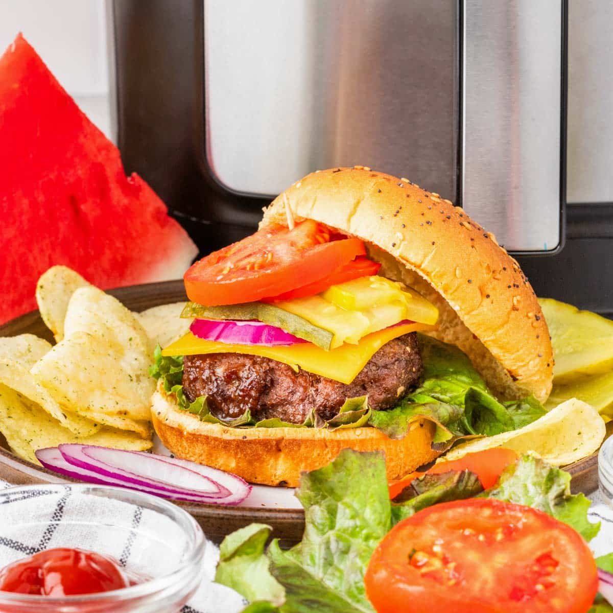 A hamburger on a bun with condiments on a plate along with chip and an air fryer in the background.