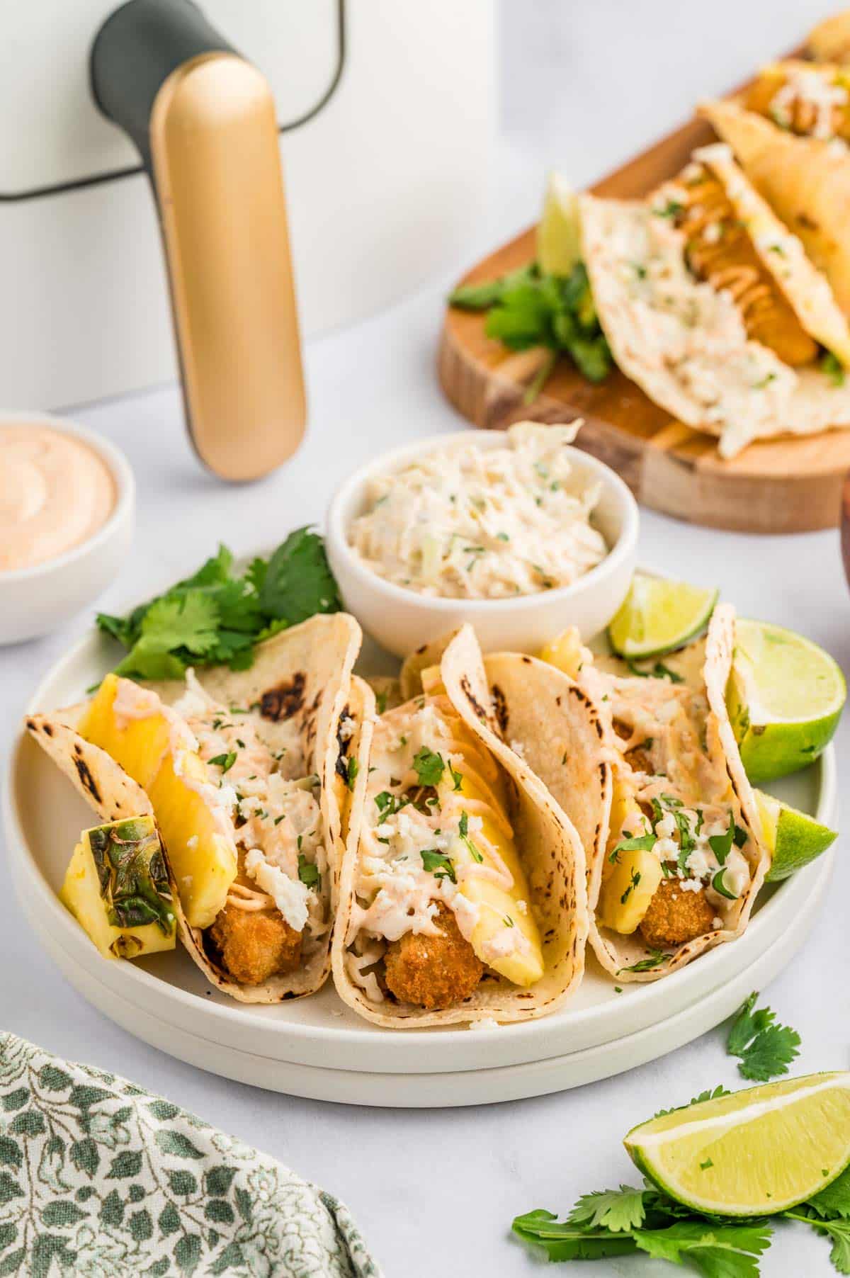 Fish tacos on a white plate, garnished with lime, pineapple, slaw, and cilantro.