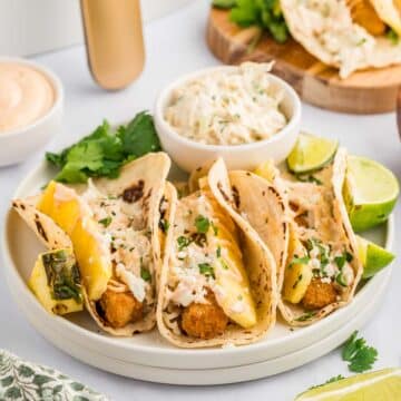 Fish tacos on a white plate, garnished with lime, pineapple, slaw, and cilantro.