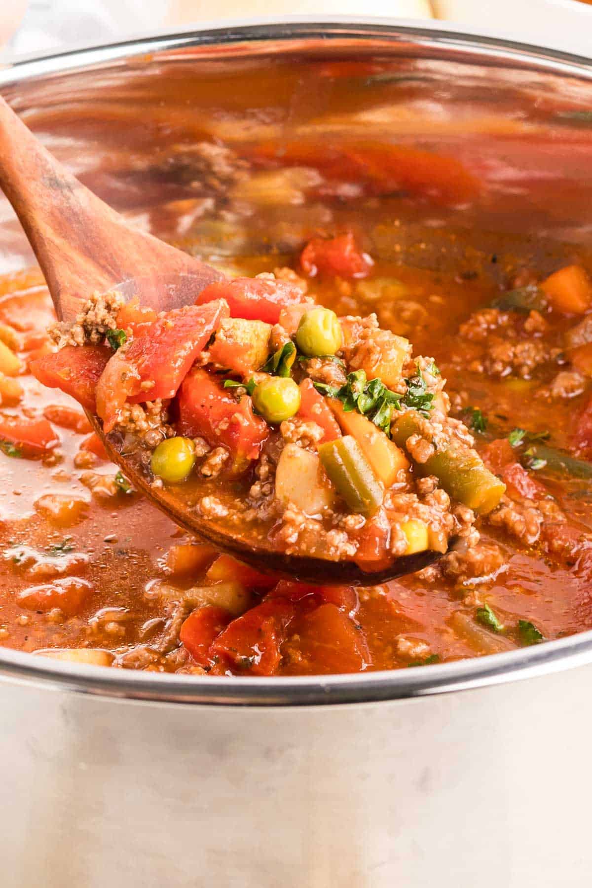 A soup pot with a wooden spoon, scooping out a portion of vegetable soup with ground beef.