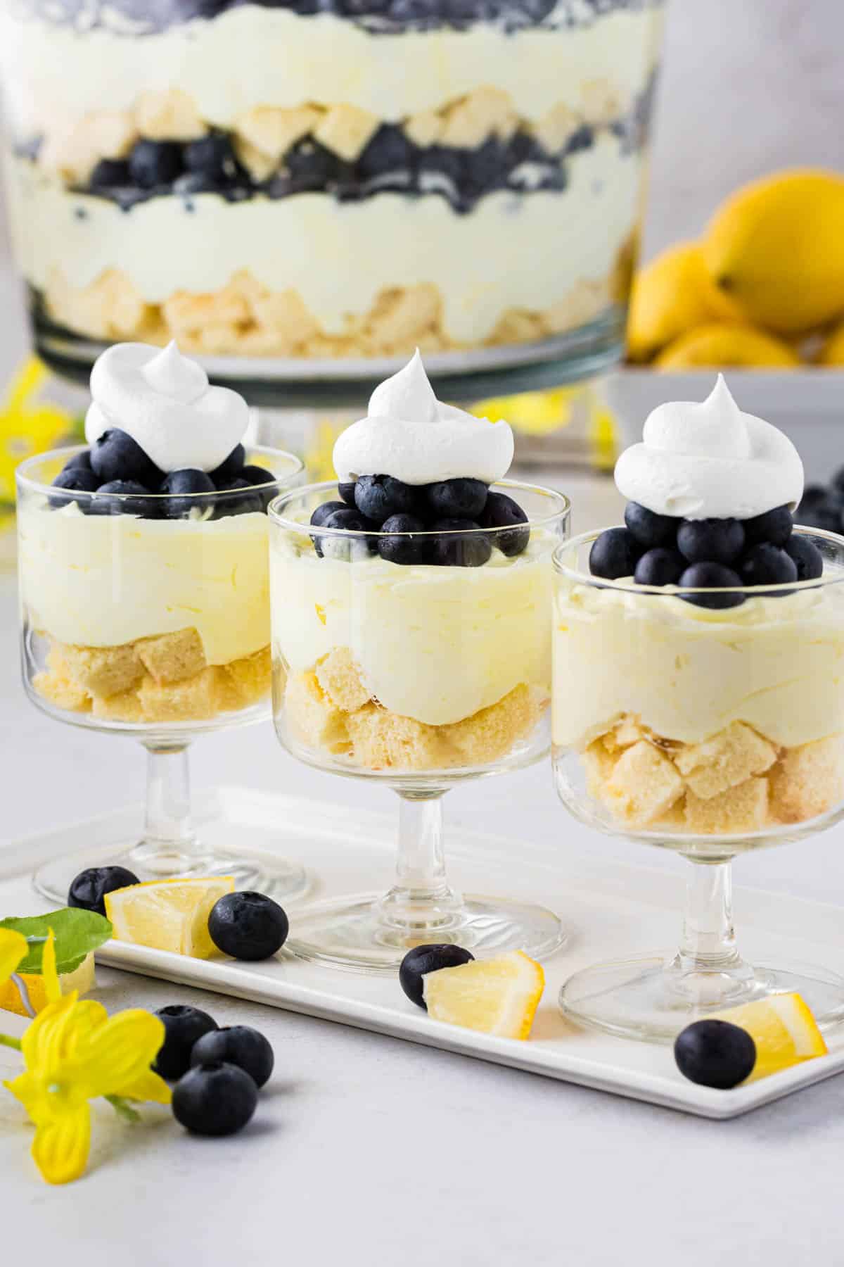 Three lemon blueberry trifles in individual stem dessert glasses with the whole trifle bowl in the background.