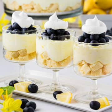 Three lemon blueberry trifles in individual stem dessert glasses with the whole trifle bowl in the background.