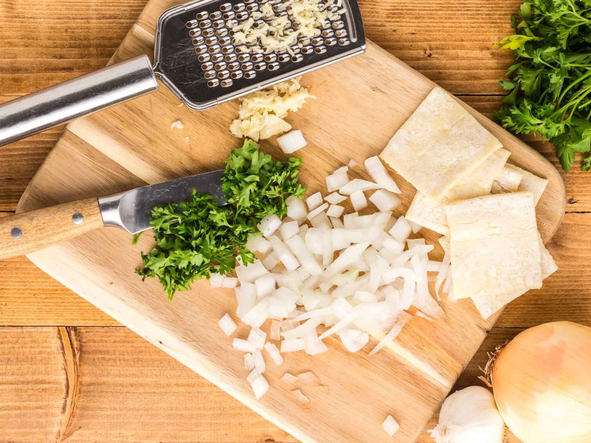 A cutting board with a knife that has chopped onions, parsley, sliced puff pastry, squares, and a greater that minced garlic for the recipe.