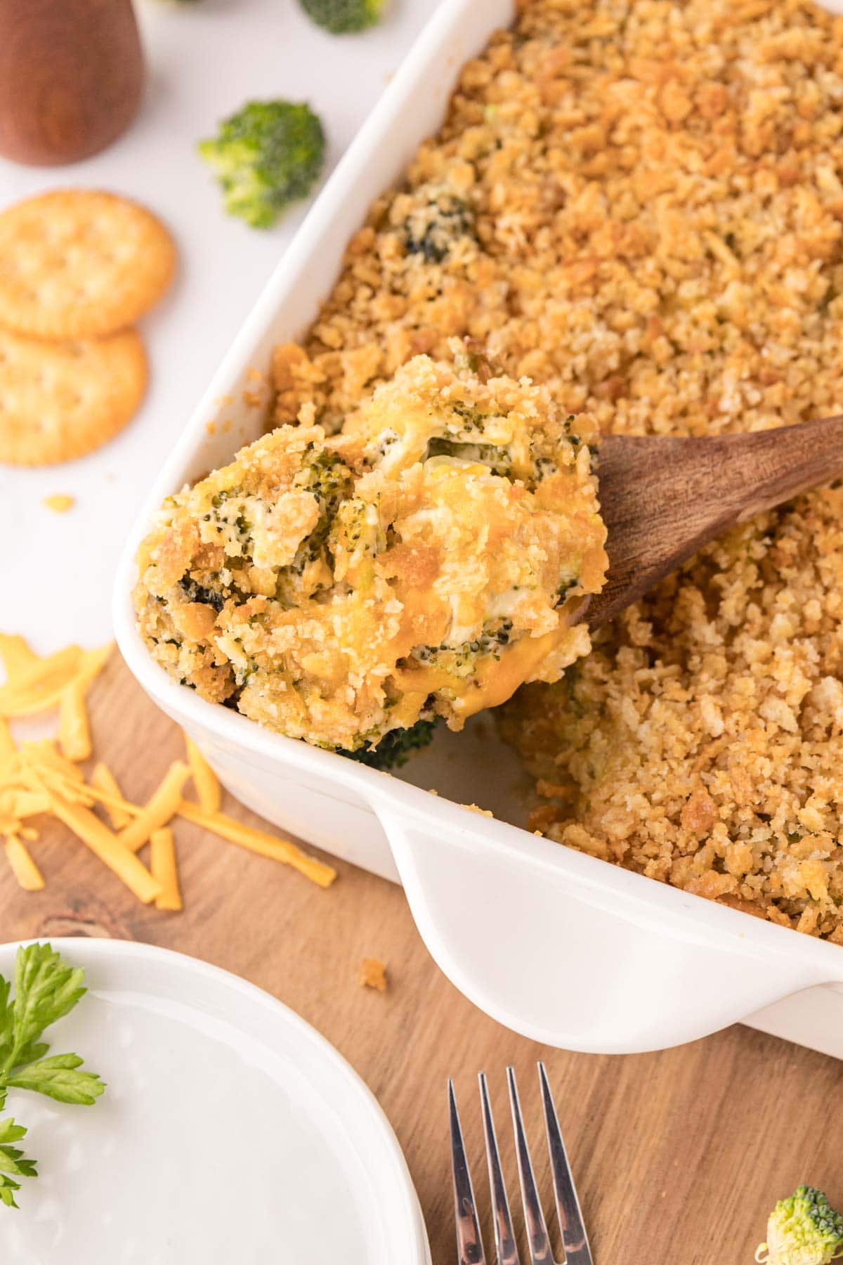 Scooping a serving of cheesy broccoli casserole with a wooden spoon from a white casserole dish.