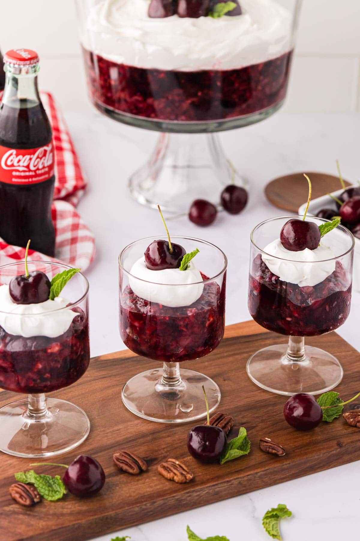 Individual cherry jello salad cups topped with whipped cream and a stemmed cherry with the  trifle bowl in the background.