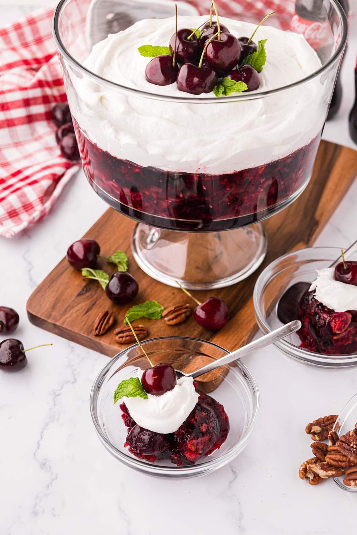 A clear serving bowl with cherry cola Jell-O salad garnished with cool whip and a mint leaf, with the trifle bowl in the background.