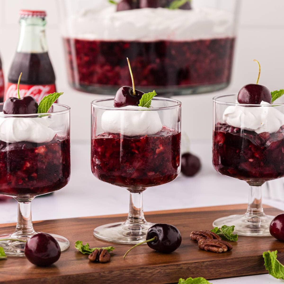 Individual cherry jello salad cups topped with whipped cream and a stemmed cherry with the trifle bowl in the background.