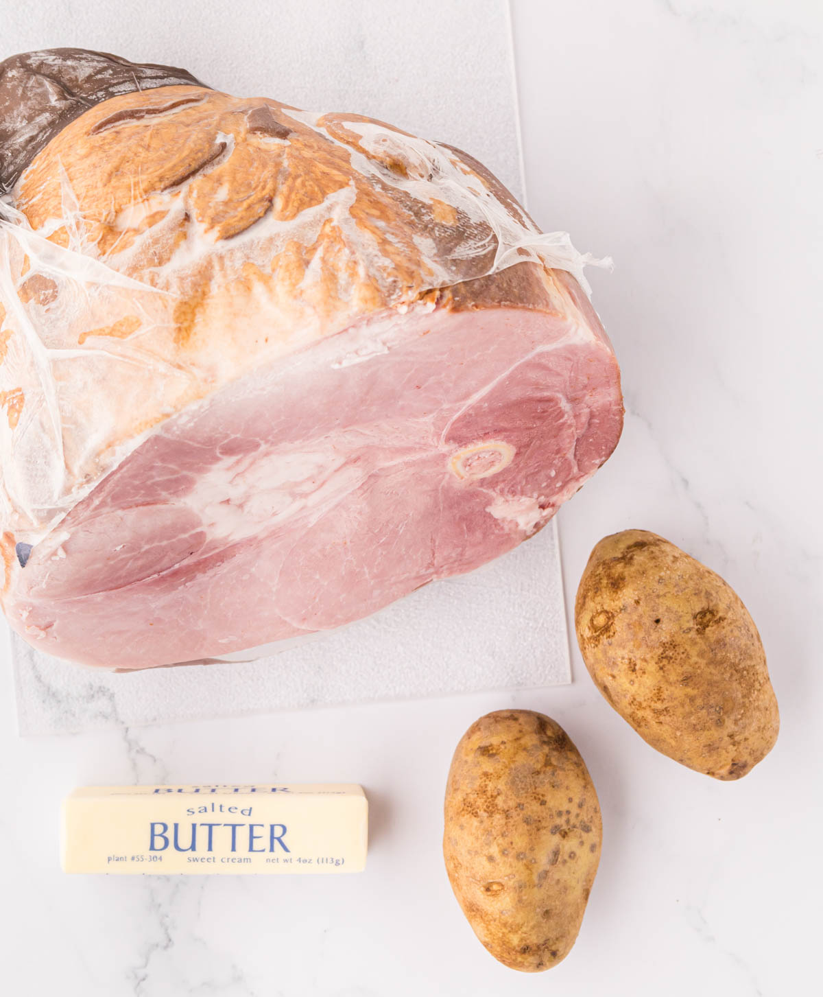 A large ham, russet potatoes, and stick of butter on a marble slab.