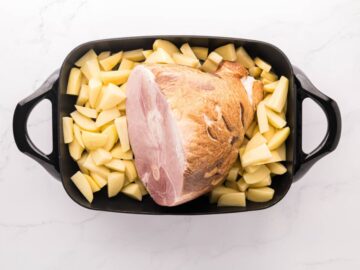 An electric skillet filled with an ucooked half ham and russet potatoes wedges.