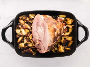 An electric skillet filled with a cooked half ham and russet potatoes wedges.