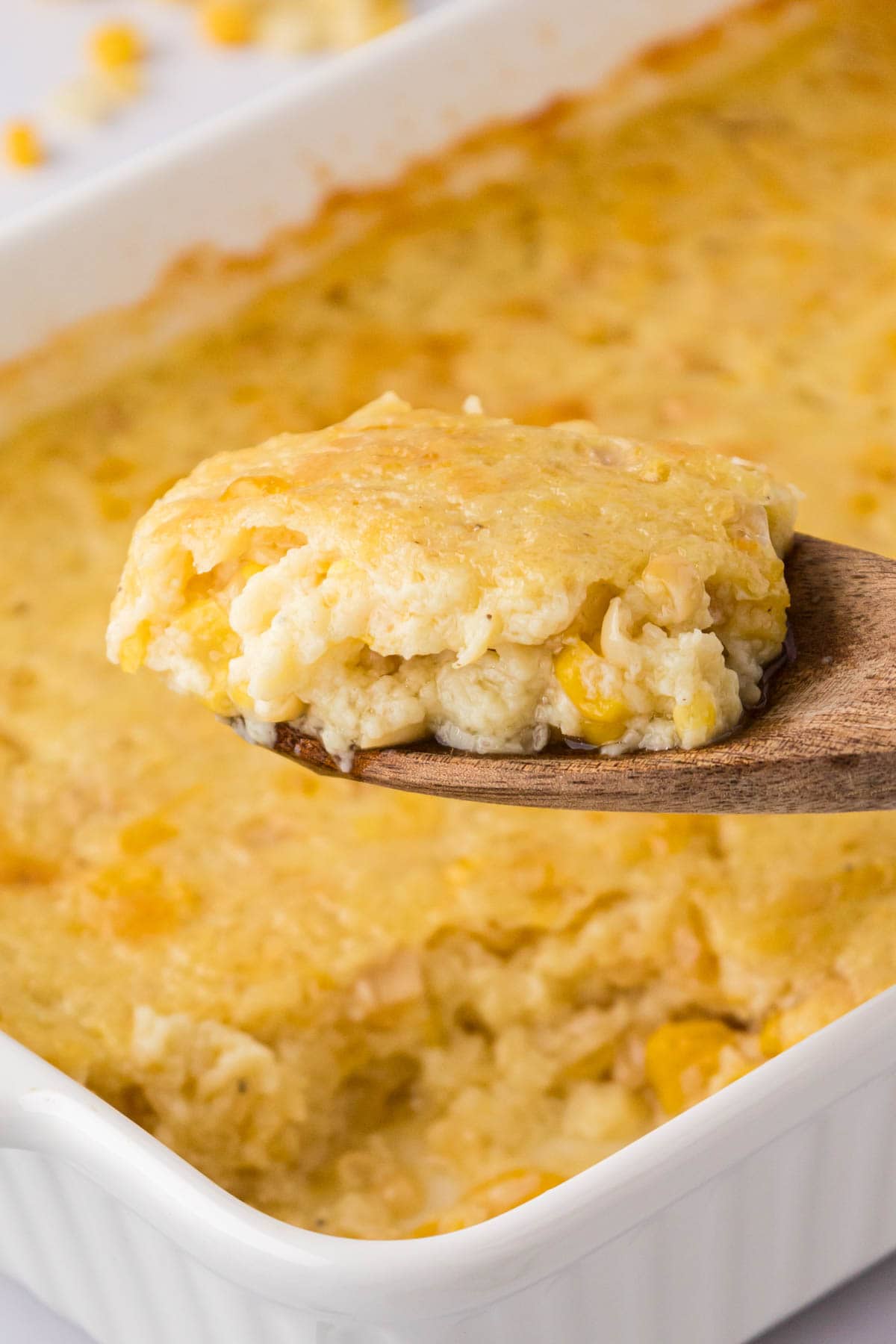 A wooden spoon, removing a serving of corn pudding from the baking dish.