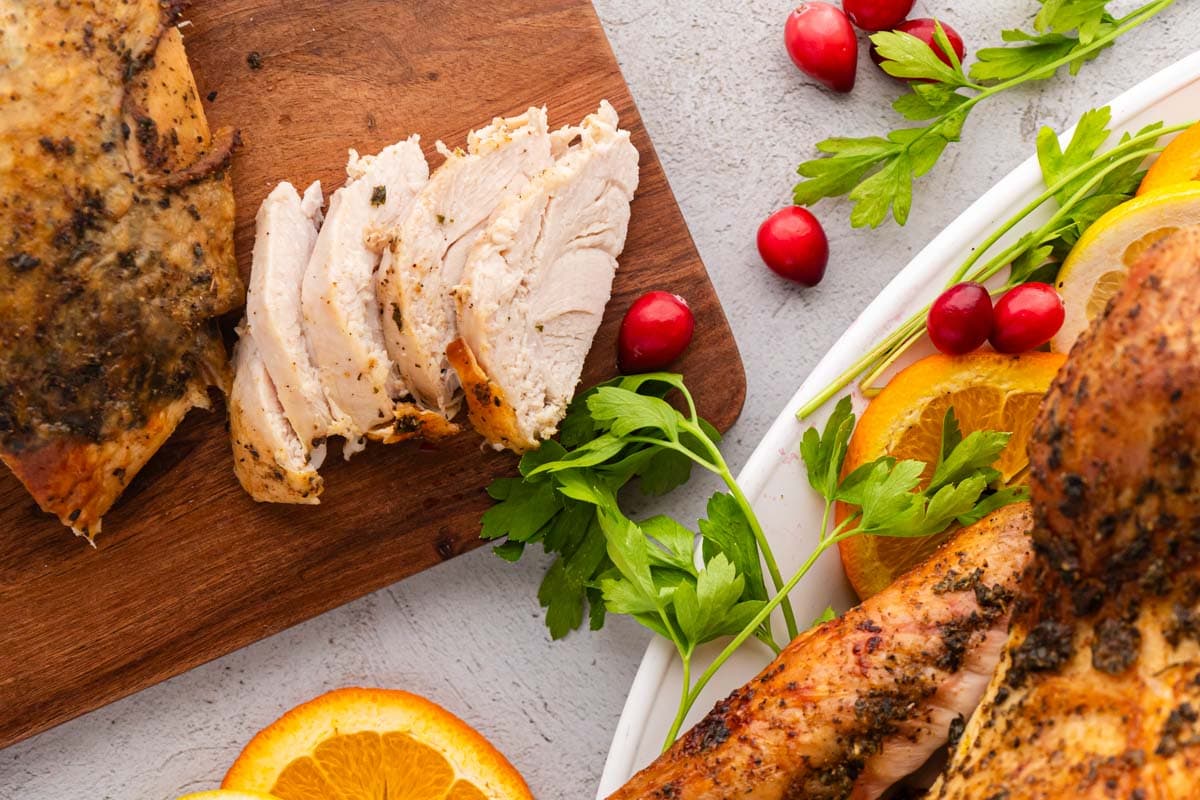 A wooden carving board with sliced turkey breast and the whole turkey st to the side on the platter.