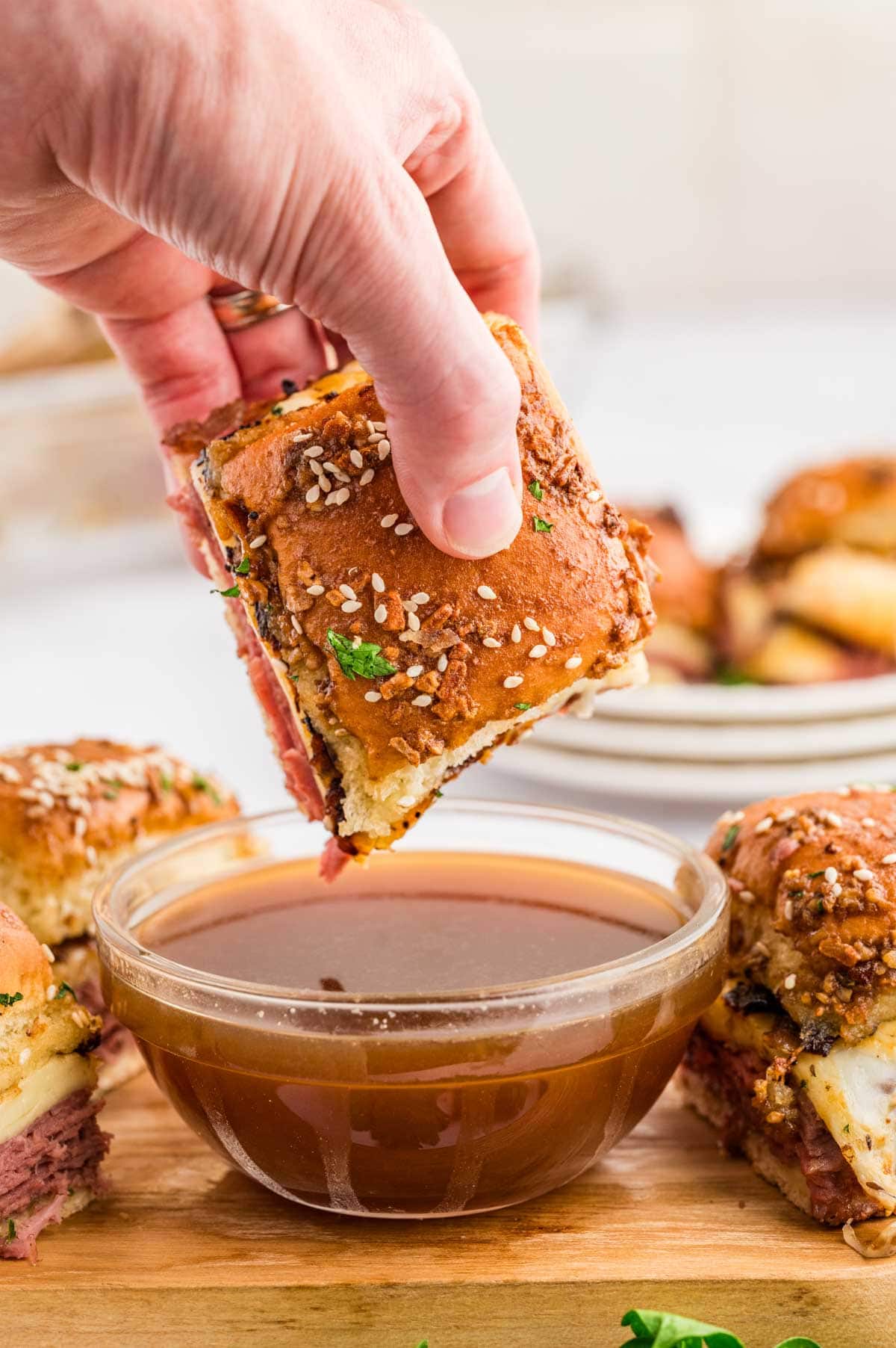 Hand dipping a French dip slider into an au jus sauce on a wooden board.