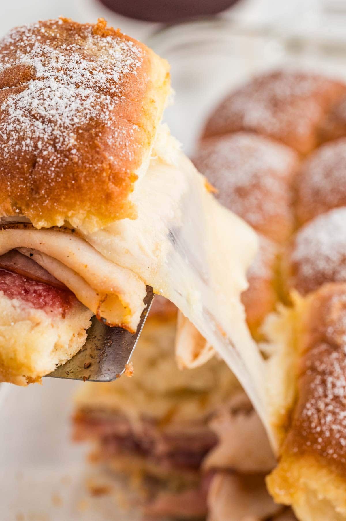 Metal spatula taking a monte criso slider from a baking dish with the cheese pulling from the rest of the sliders.