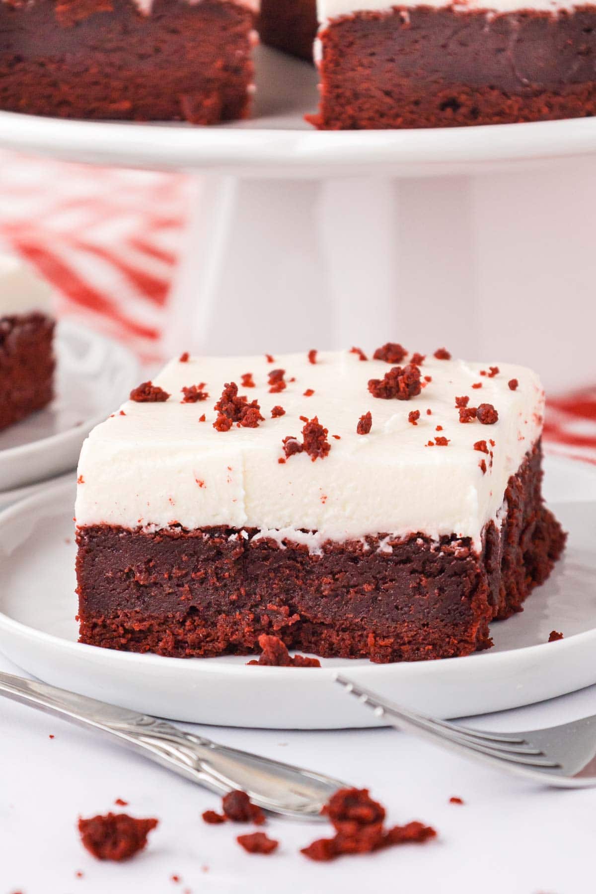 A red velvet brownie on a white plate with a cake pedestal in the background filled with brownies.