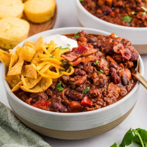 Two bowls of chili with fritos, shredded cheese, and sour cream on top and corn bread muffins in the background.