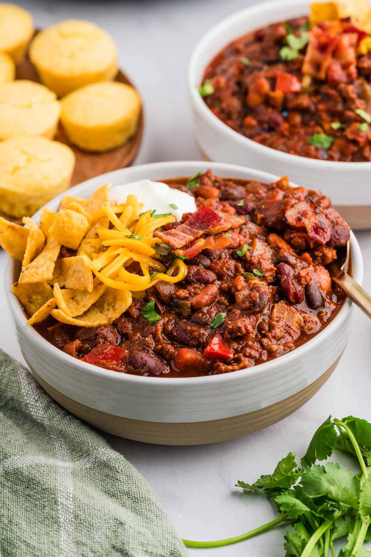 Two bowls of chili with fritos, shredded cheese, and sour cream on top and corn bread muffins in the background.