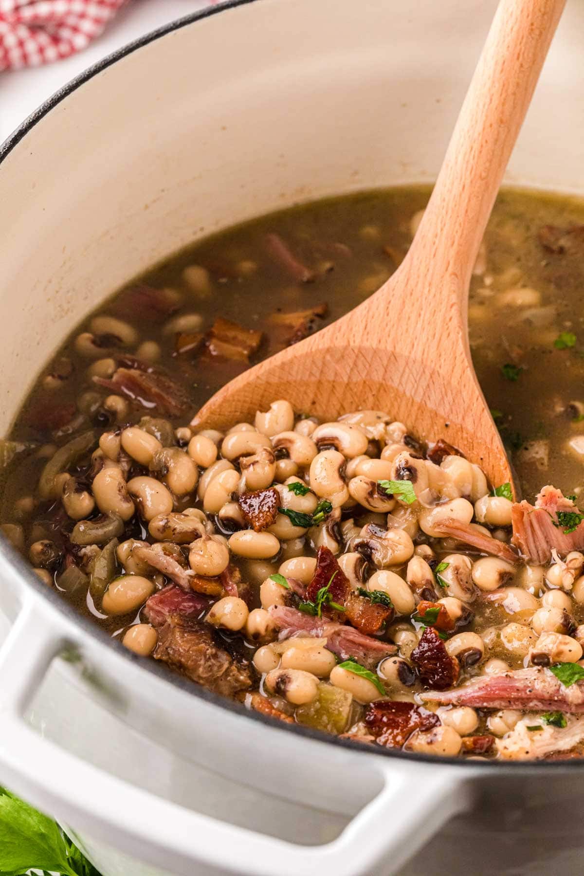 A white Dutch oven filled with southern black-eyed peas and a wooden spoon, removing a scoop of peas from the pot.