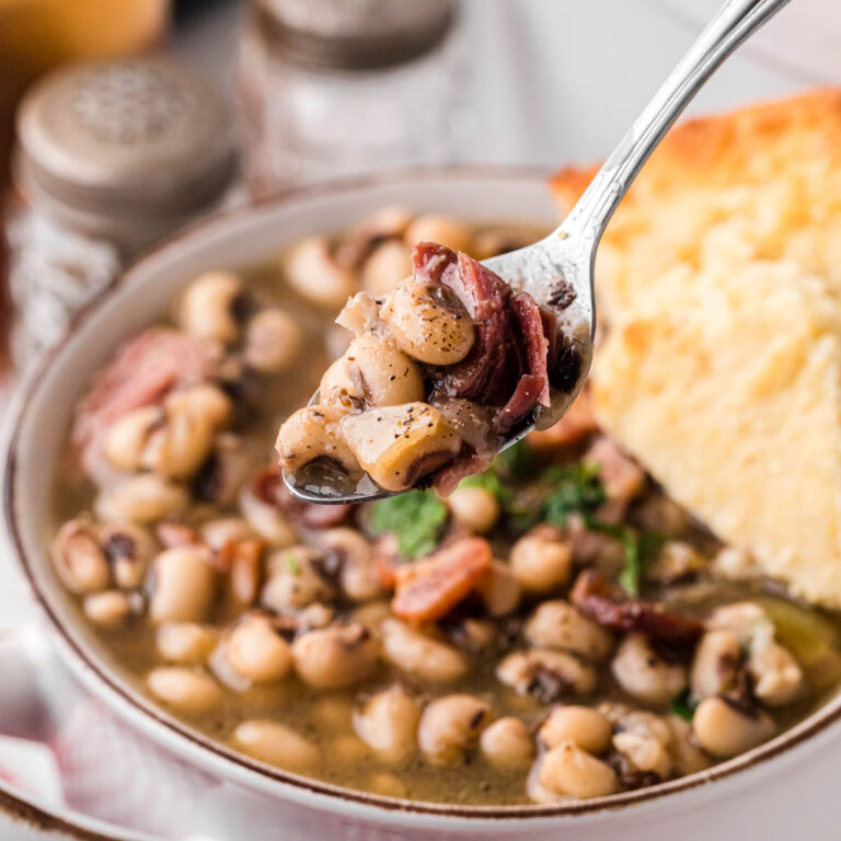 A bowl of Black Eyed Peas with a slice of cornbread and a silver spoon removing a bite of peas.