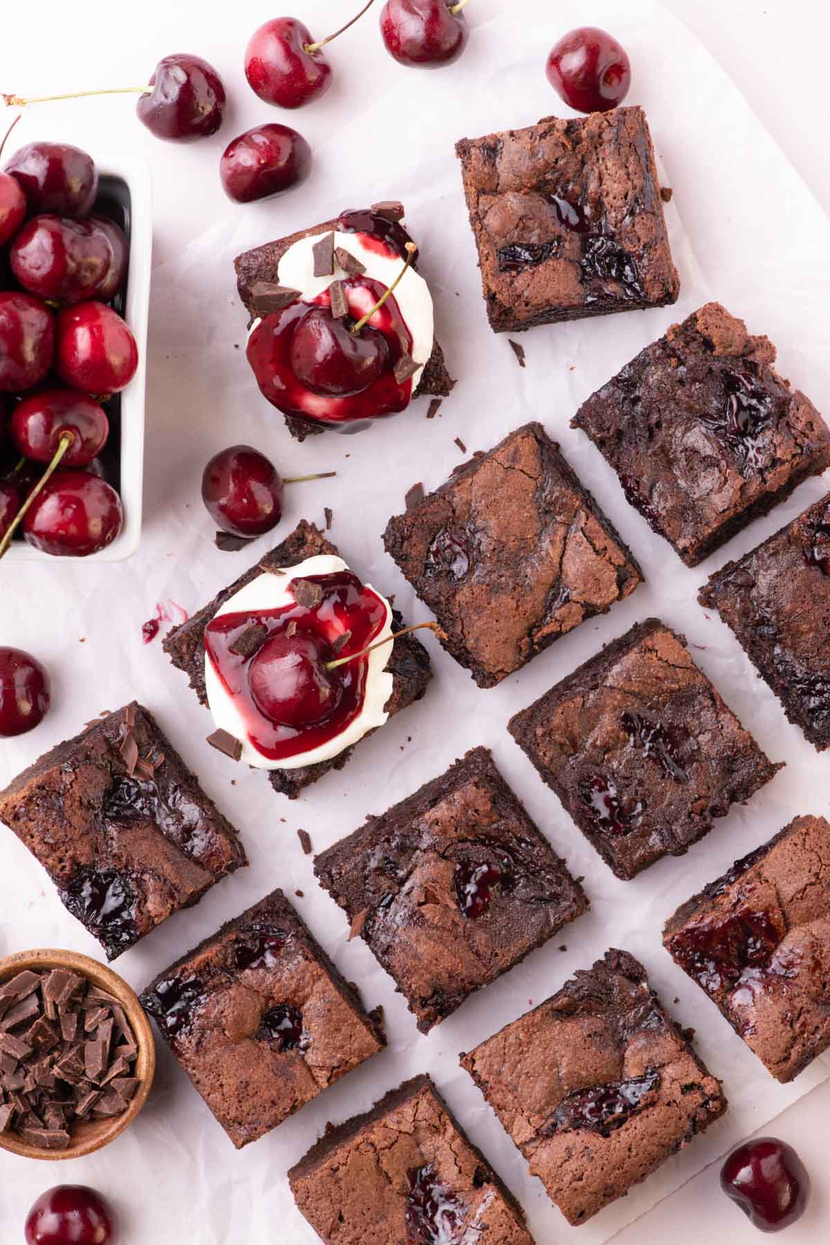 Black forest brownies on parchment with some of them topped with whipped cream and extra cherry filling.