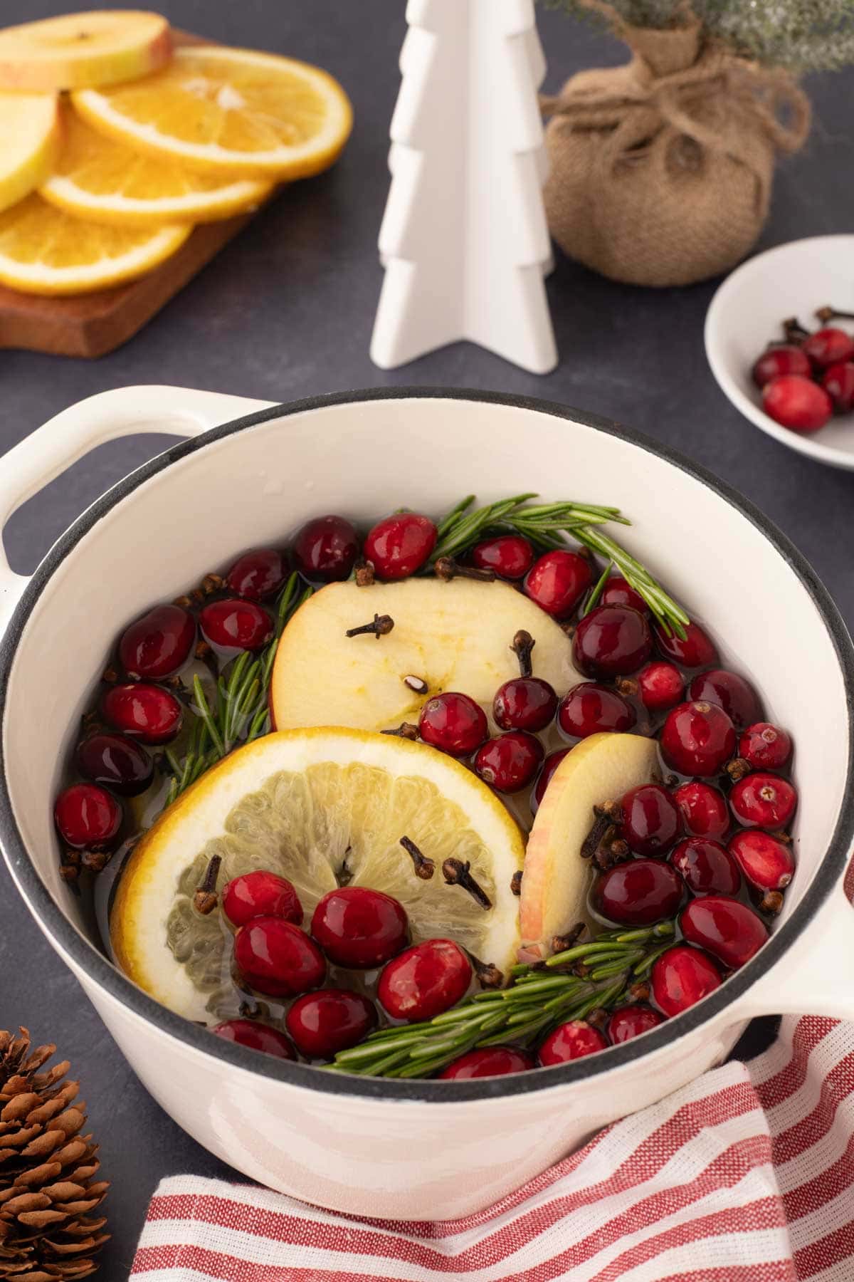 White simmer pot with a black rim with water, sliced oranges, apples, clothes, cranberries, and rosemary.