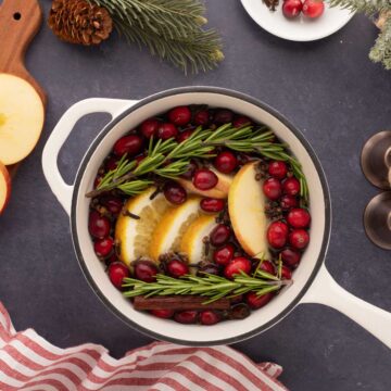 White simmer pot with a black rim with water, sliced oranges, apples, clothes, cranberries, and rosemary.