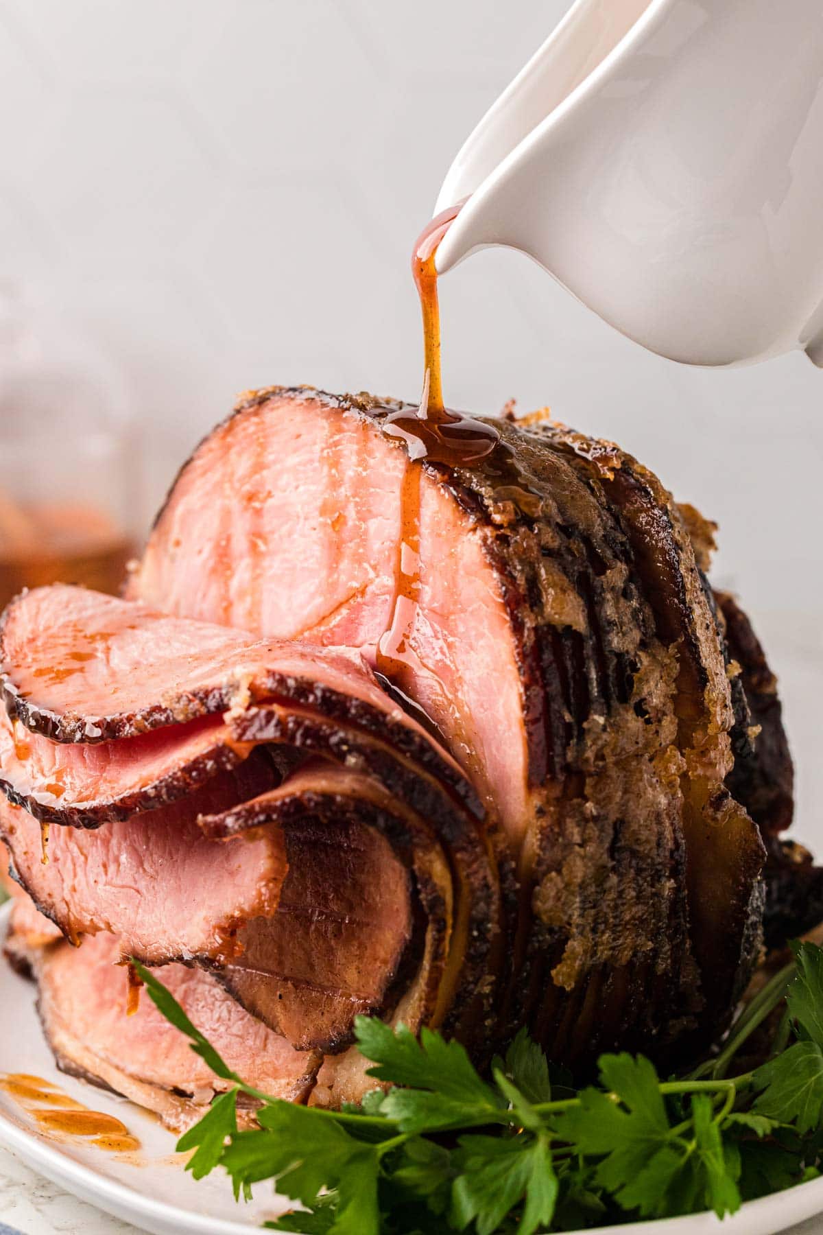 A white serving plate with a sugar spice crusted honeybaked ham with a honey glaze sauce being poured over the ham.