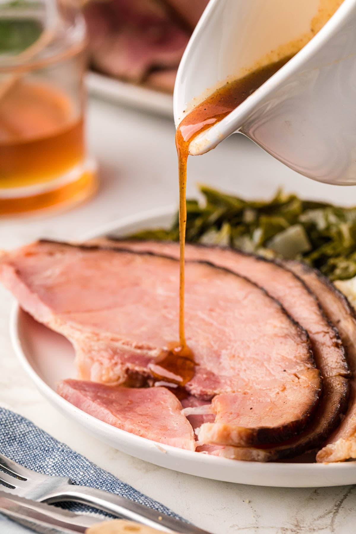 A plate of sliced ham collard greens with a glaze being poured over the ham from a gravy boat.