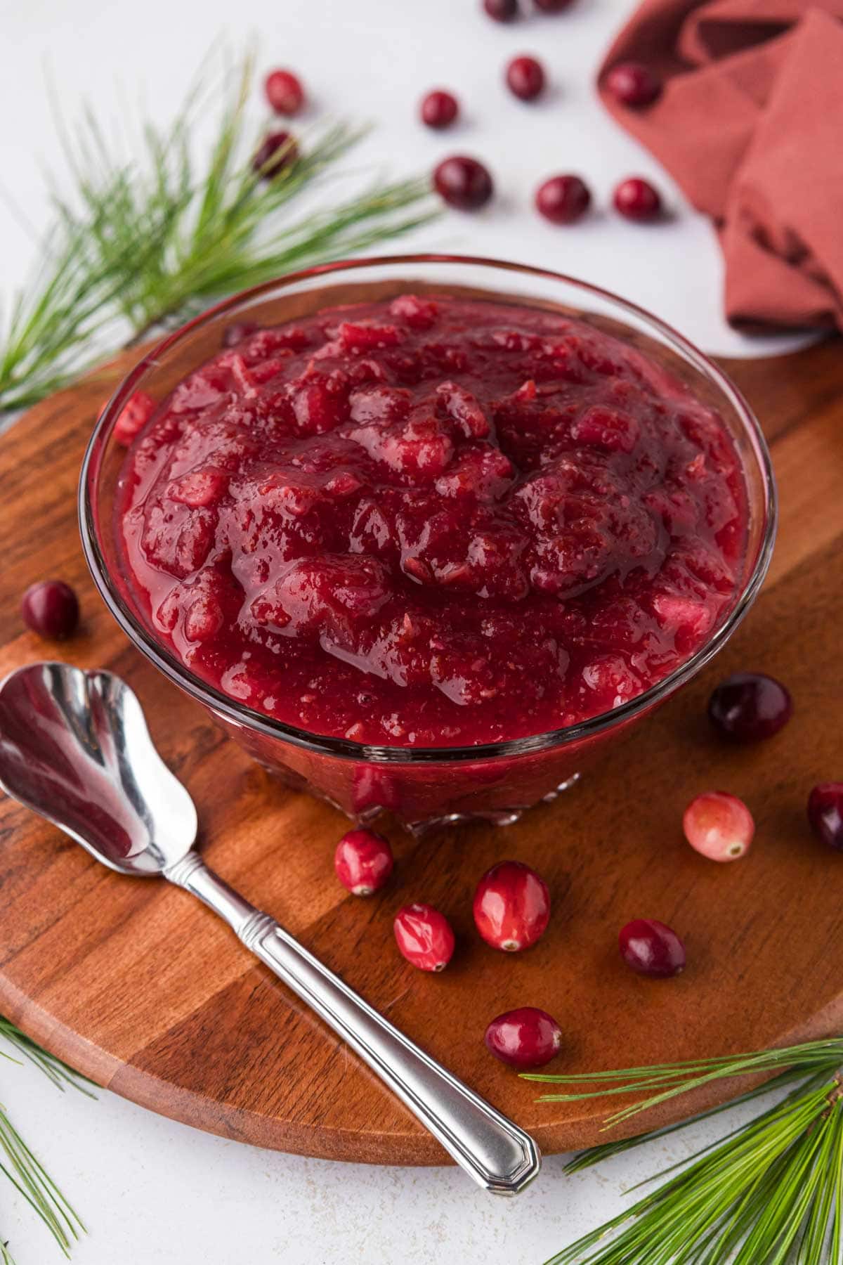 A glass bowl filled with cranberry sauce set on a round wooden board with a silver relish spoon.