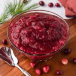 A glass bowl filled with cranberry sauce set on a round wooden board with a silver relish spoon.