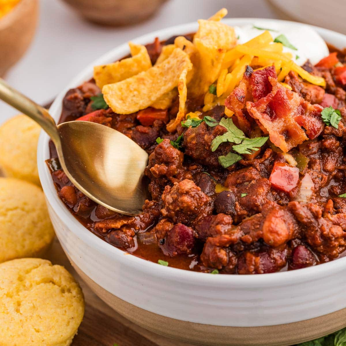 A bowl of chili with Fritos, shredded cheese, and sour cream on top and corn bread muffins in the background.