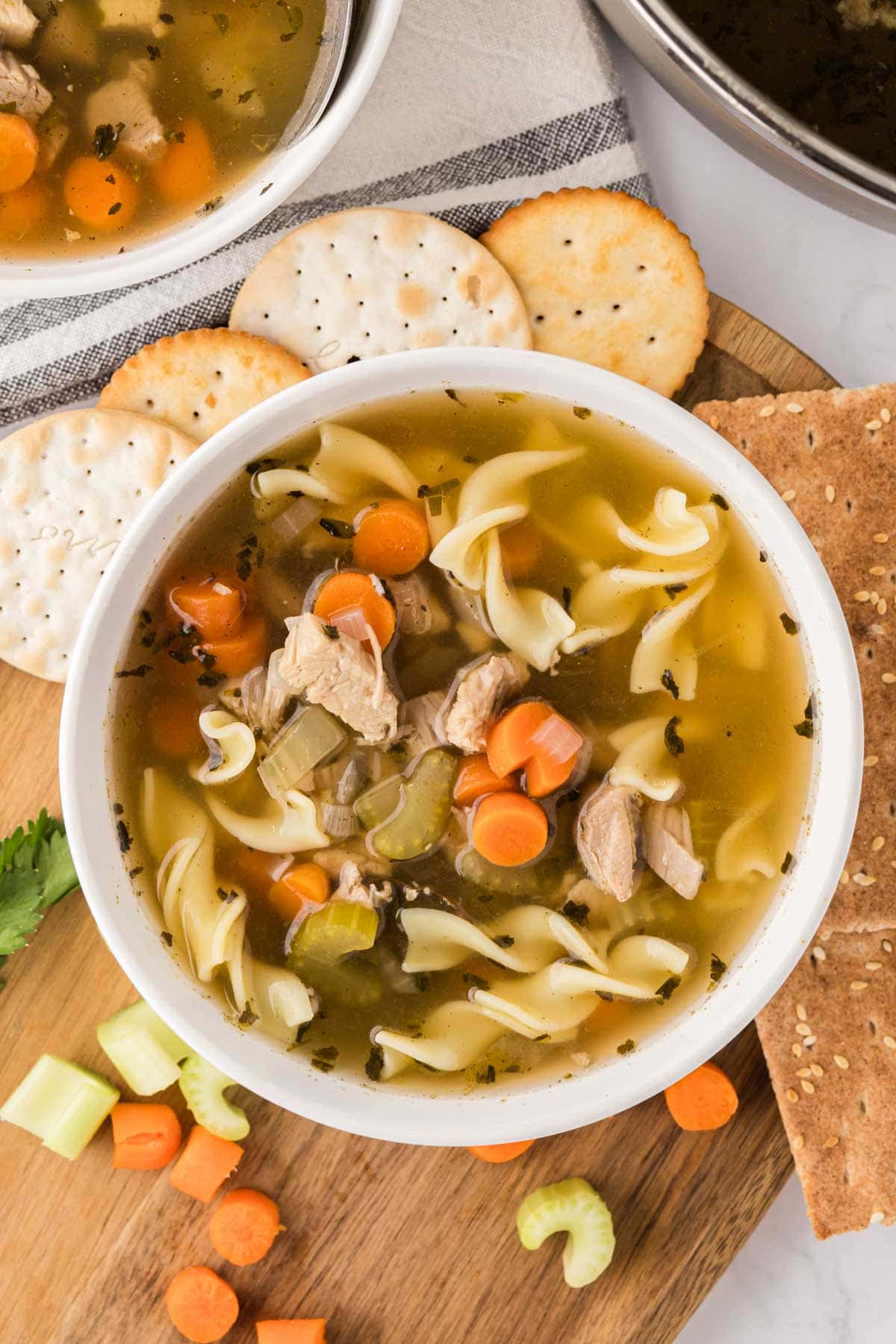 A bowl of turkey noodle soup on a wooden board served with crackers.