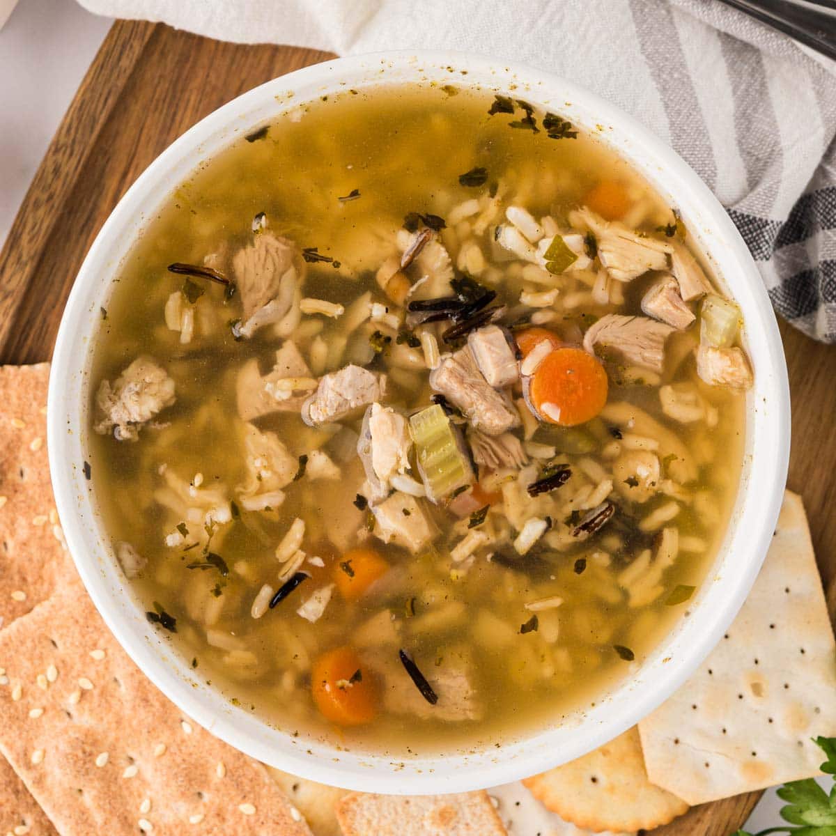 A bowl of turkey soup with rice on a wooden board served with crackers.