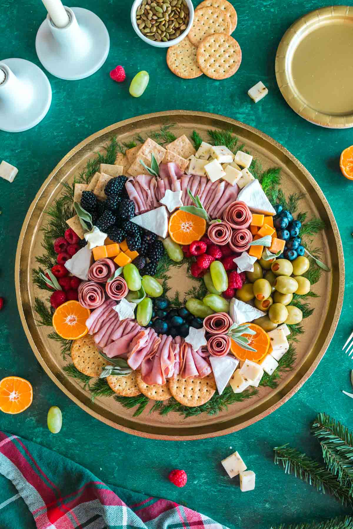 A wreath shaped charcuterie board set on a green tablecloth decorated for Christmas.