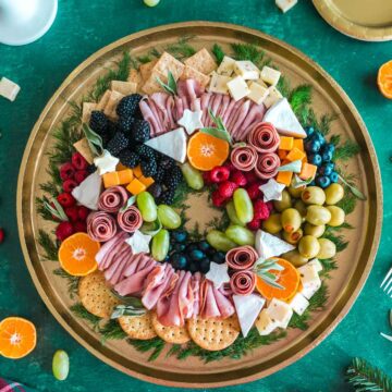 A wreath shaped charcuterie board set on a green tablecloth decorated for Christmas.