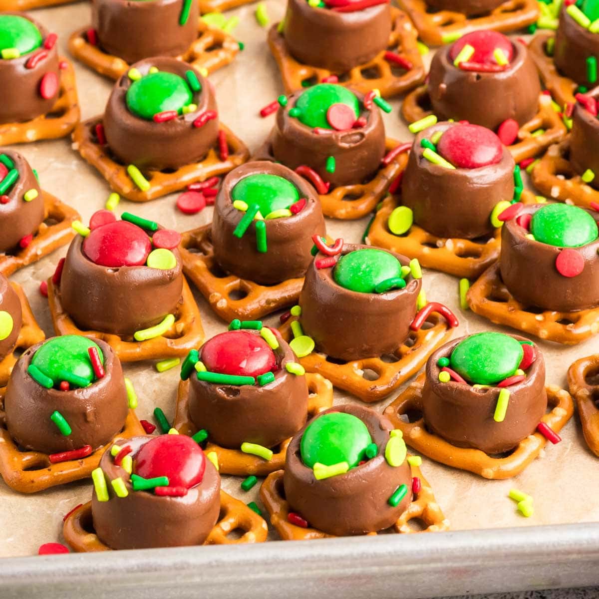 Red and green M&Ms and festive sprinkles on top of a Rolo candy that is slightly melted on top of square shaped pretzels.