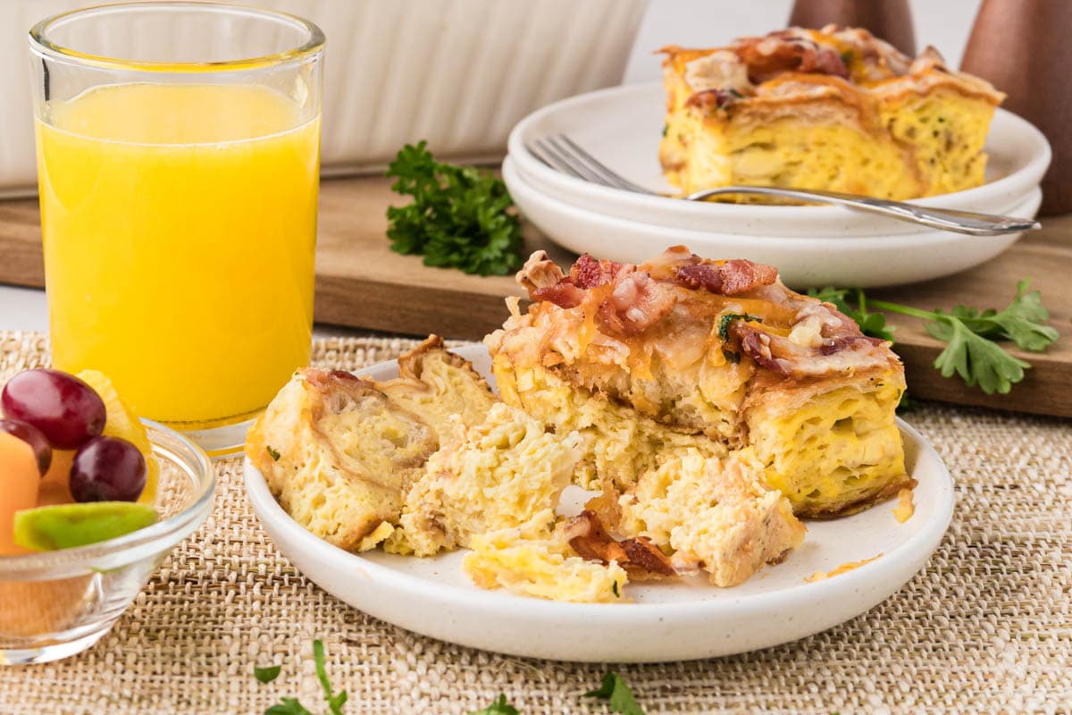 A table setting with the casserole on a plate, sliced with orange juice in the background and a bowl of fruit to the side.