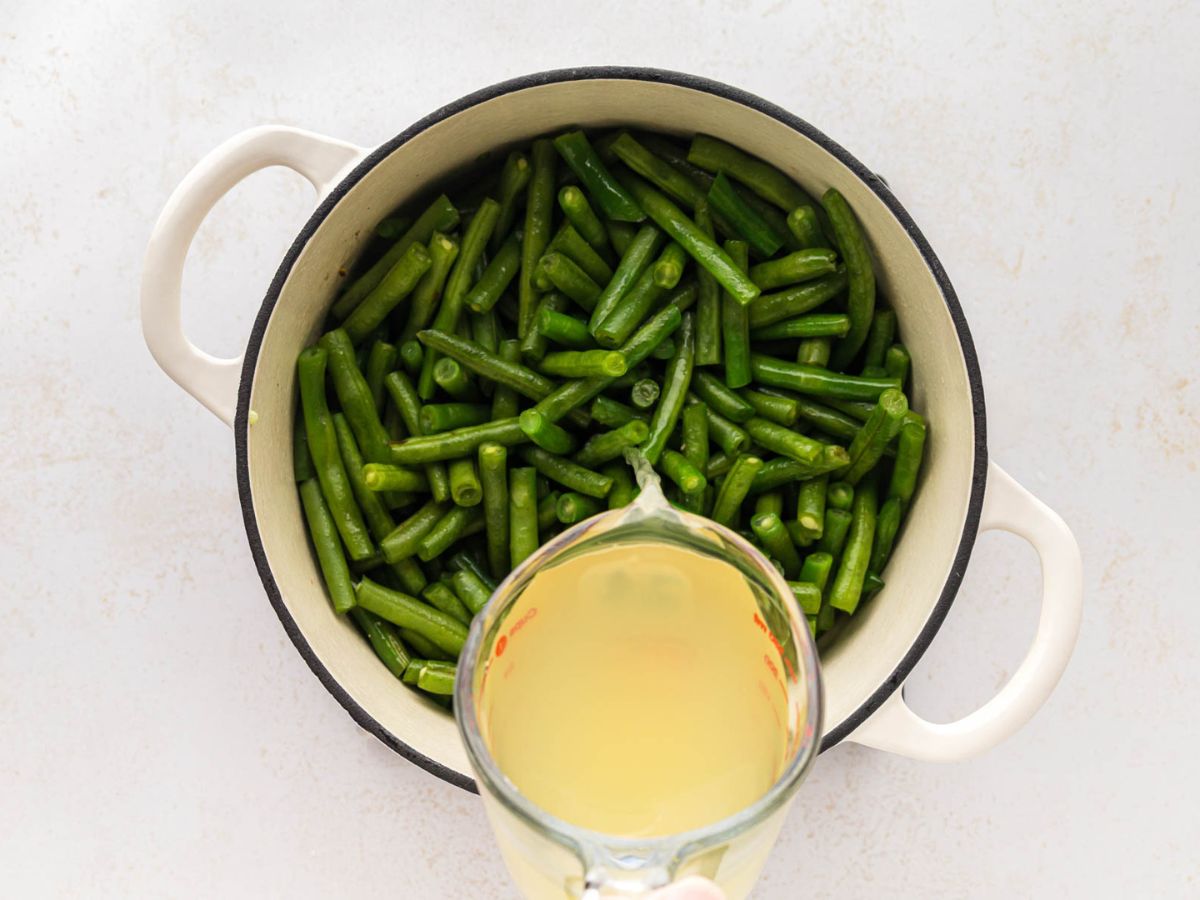 The pot full of green beans with chicken broth being poured into the pot.